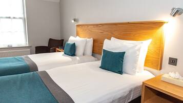 Quarto de hotel com duas camas, cada uma com roupa de cama branca e almofadas verde-água. Possui cabeceira de madeira, mesa de cabeceira com abajur e janela com persianas.