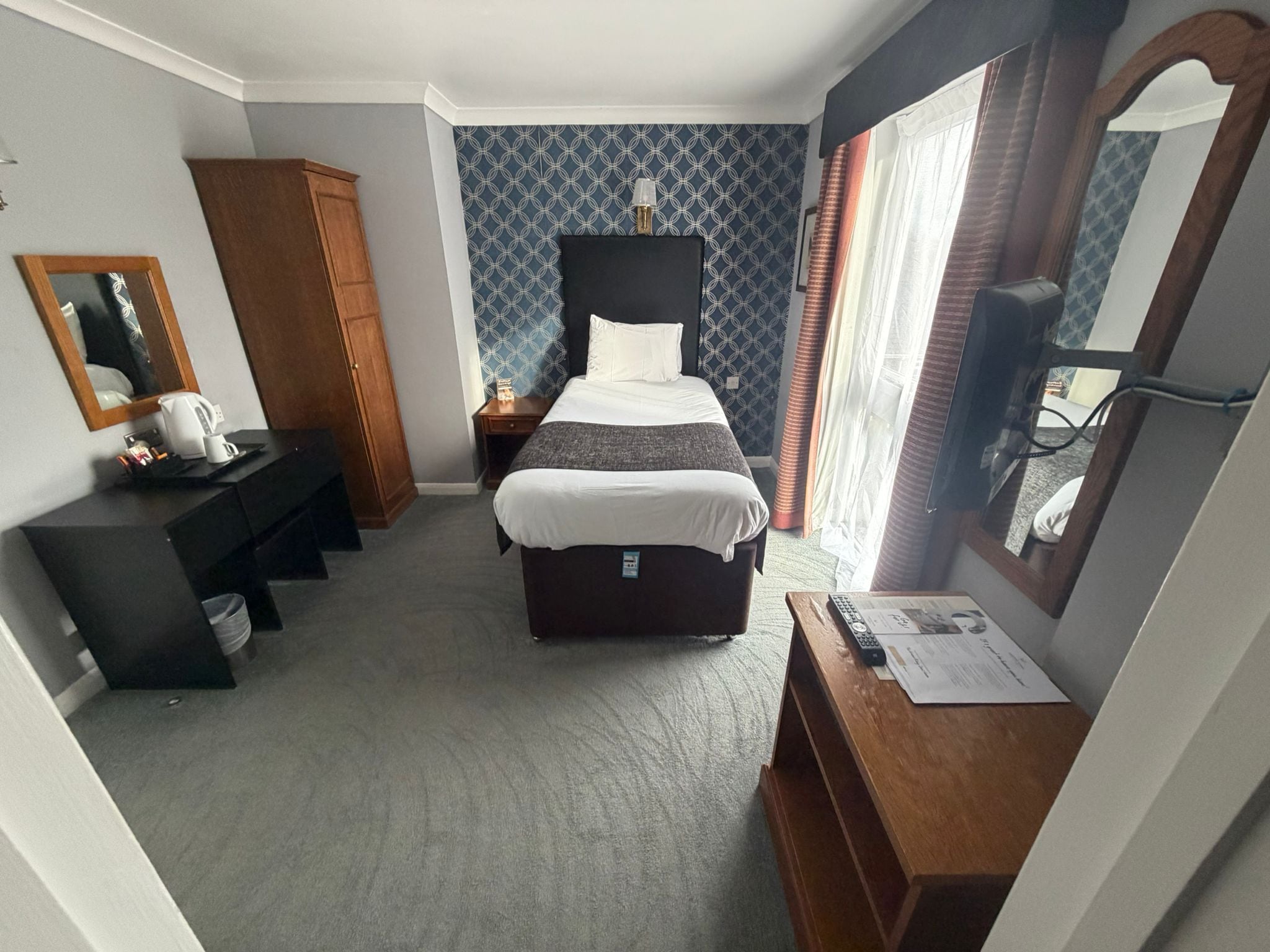 A hotel room with a bed, a desk, a wardrobe, a mirror, and a window with curtains.