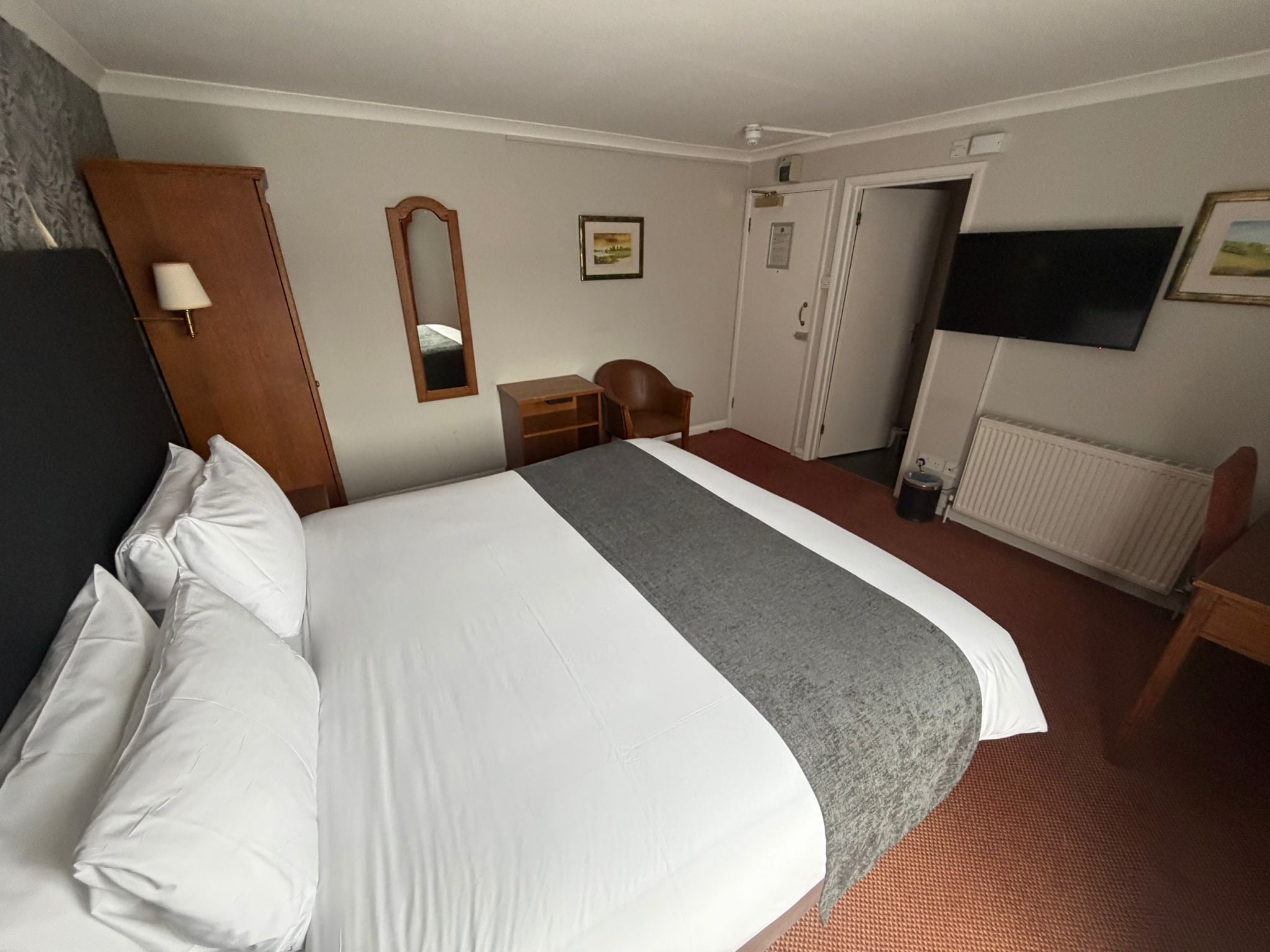 A hotel room with a large bed, a desk, a chair, a TV, and a wardrobe.