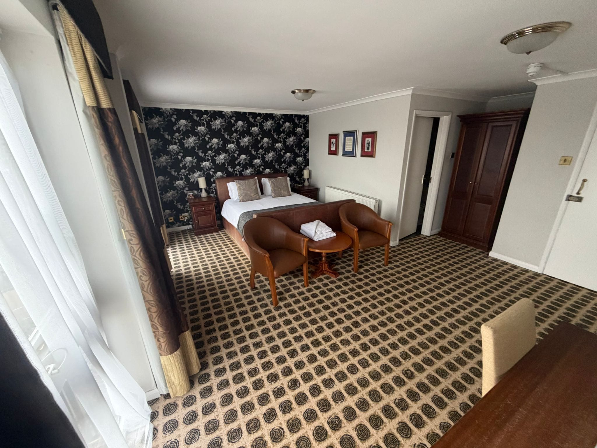 Habitación de hotel con cama, escritorio, silla, alfombra estampada, papel tapiz con diseño floral oscuro y una puerta.