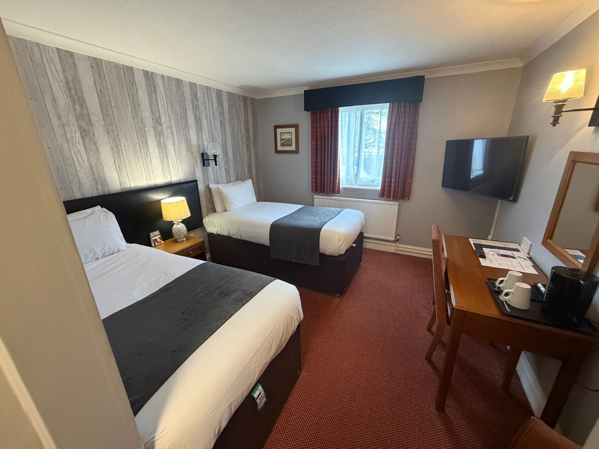 A hotel room with two beds, a desk with a chair, a TV, and a window with curtains.