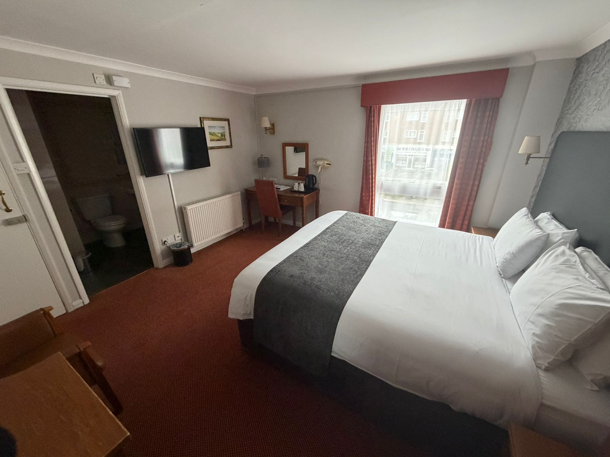 Habitación de hotel con cama, escritorio, silla, televisor y ventana con cortinas.