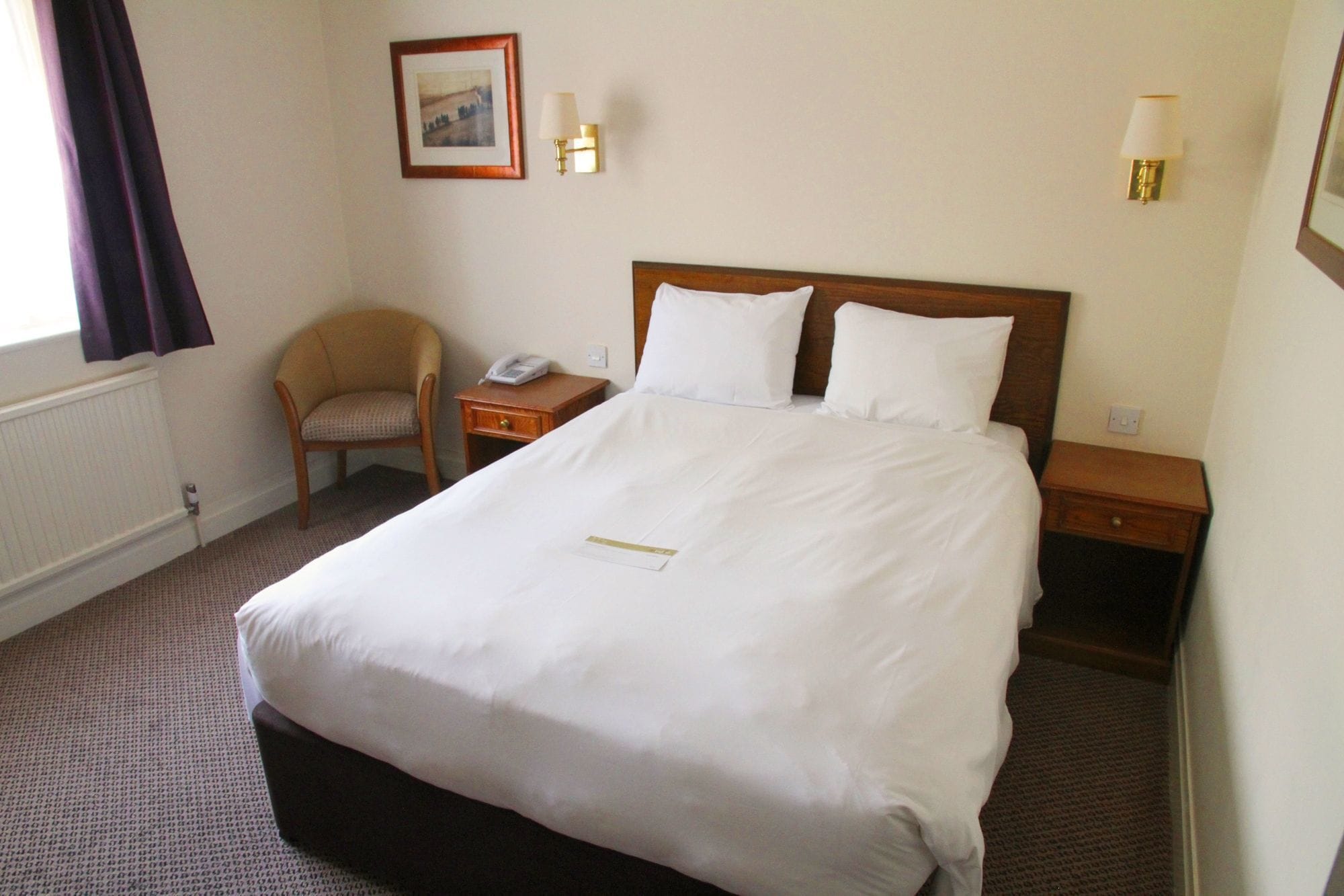 A hotel room with a large bed, a chair, a bedside table with a lamp, and a framed picture on the wall.