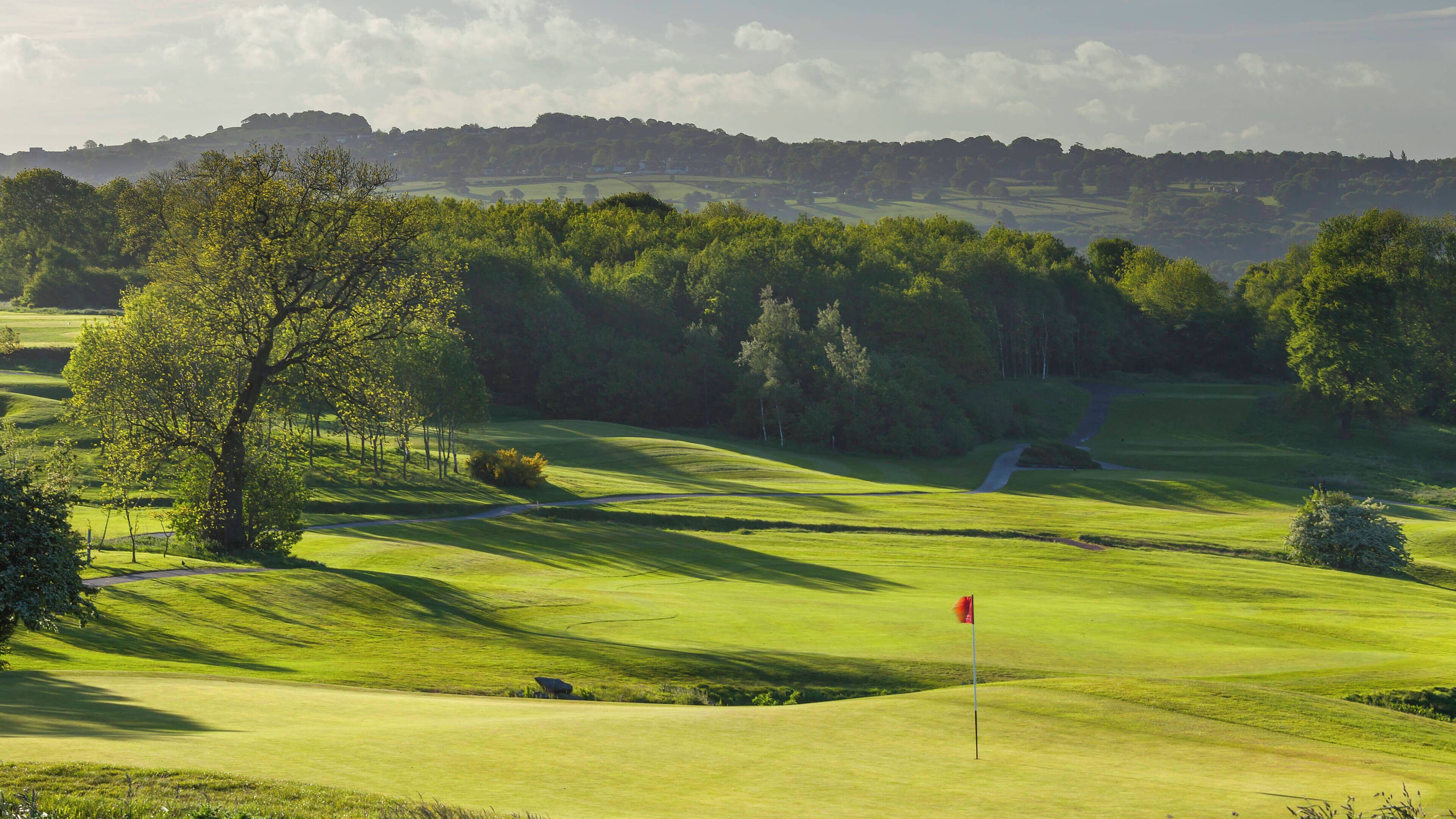 Un campo de golf con colinas verdes ondulantes, un palillo de señalización y un sendero que serpentea entre los árboles.