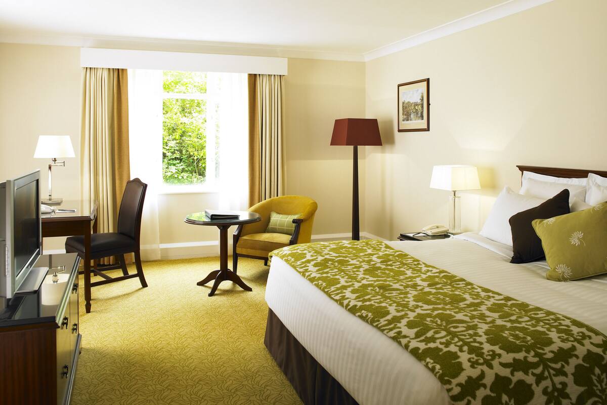 Ein Hotelzimmer mit Bett, Schreibtisch mit Fernseher, Stuhl, kleinem Tisch und einem Fenster mit Vorhängen.
