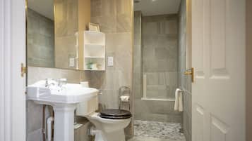 A bathroom with a white pedestal sink, a toilet with a wooden seat, a shower area, and a tiled floor.