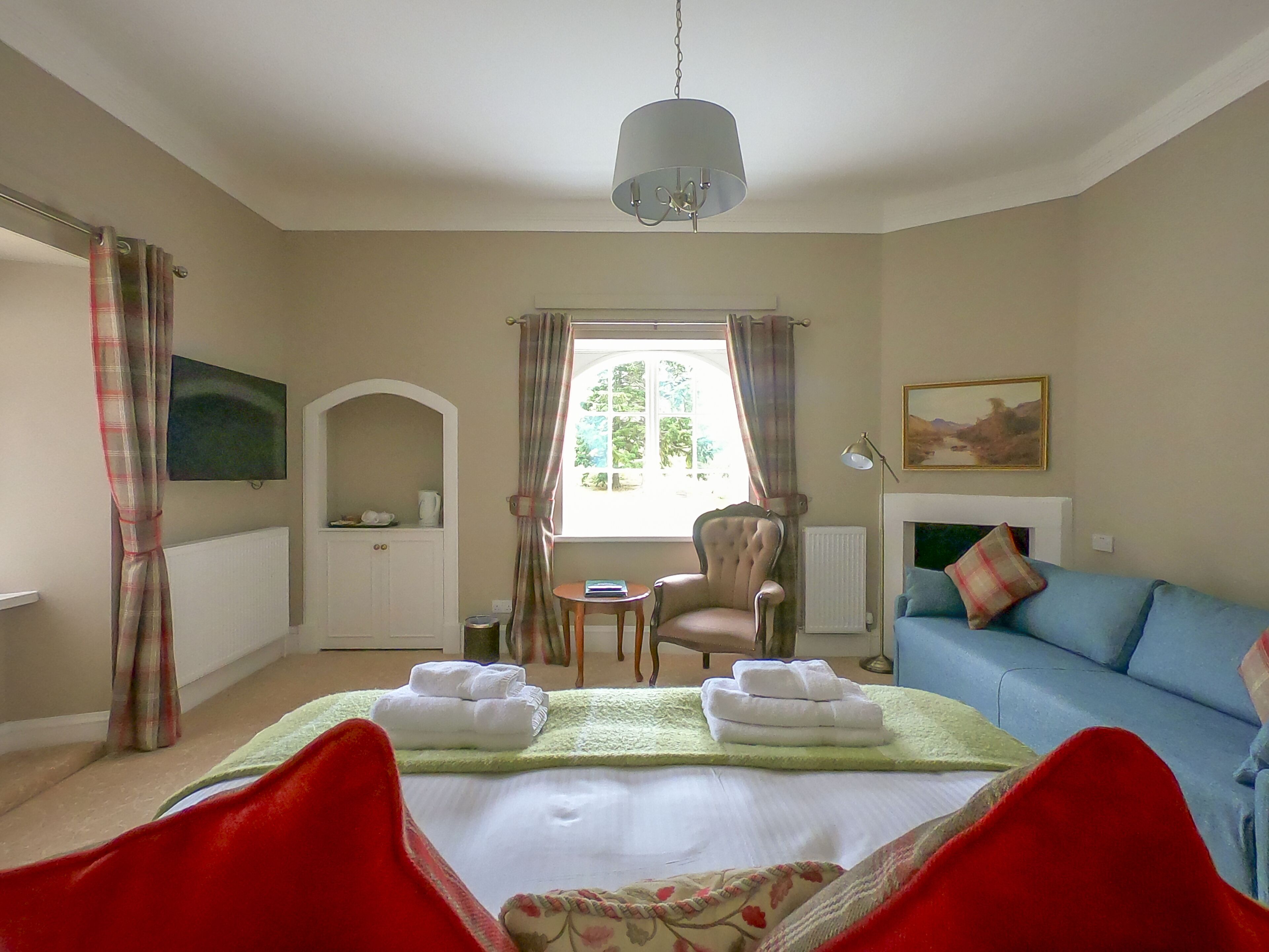 Quarto de hotel com cama, poltrona, sofá, televisão, janela com cortinas e um quadro na parede.