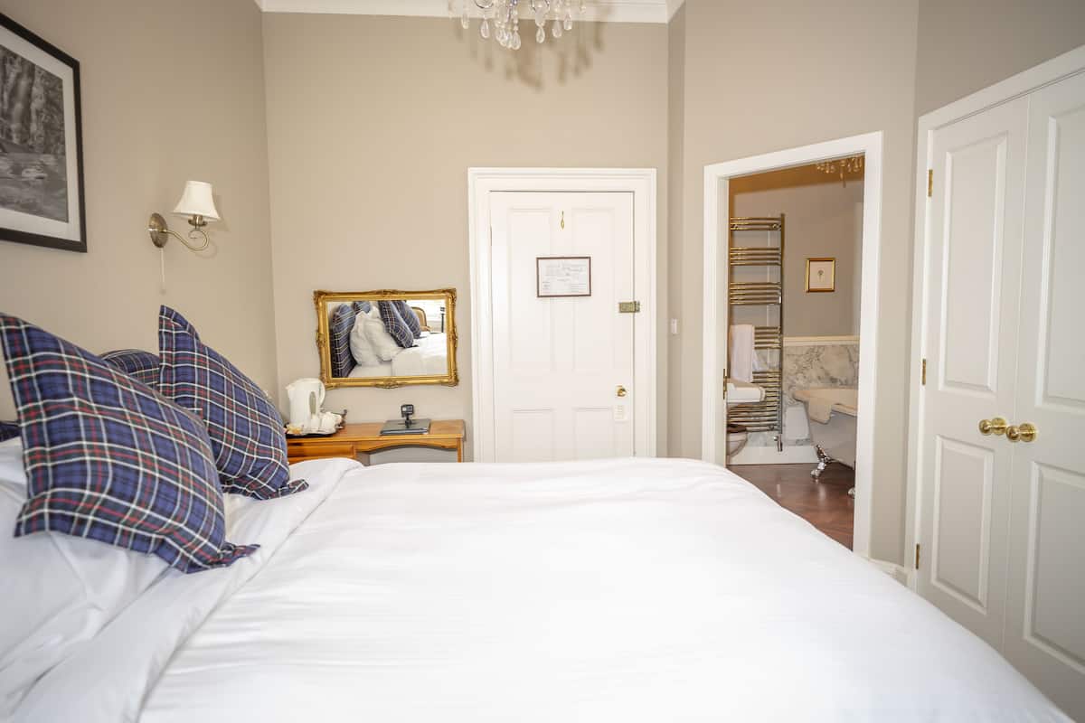 Un lit bien fait, agrémenté de coussins à carreaux, une table de chevet avec une lampe et une porte donnant sur une salle de bain équipée d’un porte-serviettes.