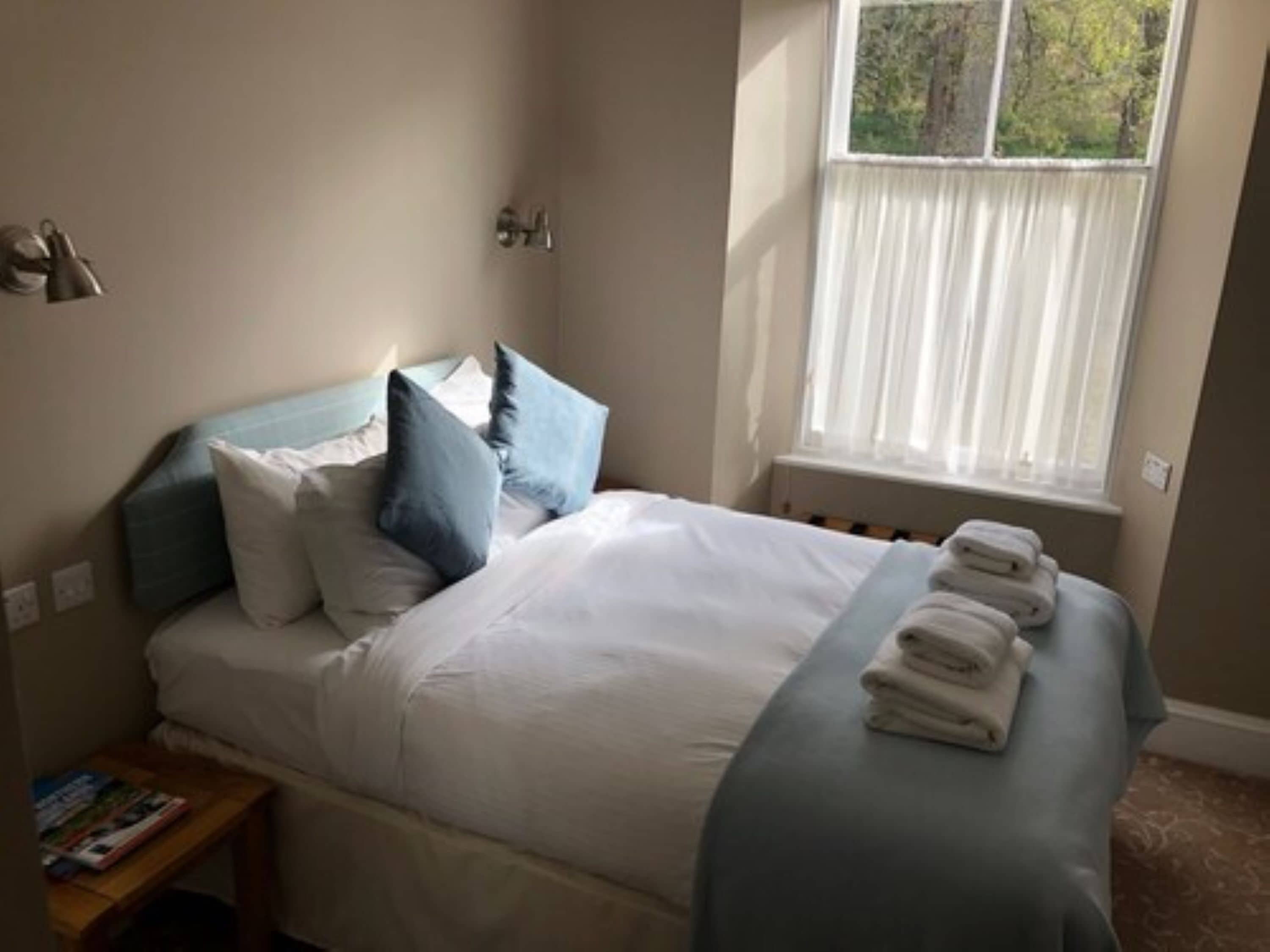 A neatly made bed with white and blue bedding, a bedside table with a book, and a window with curtains.