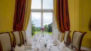 Une table à manger dressée pour un repas, avec des chaises, une vue sur un jardin par la fenêtre et des rideaux de chaque côté.