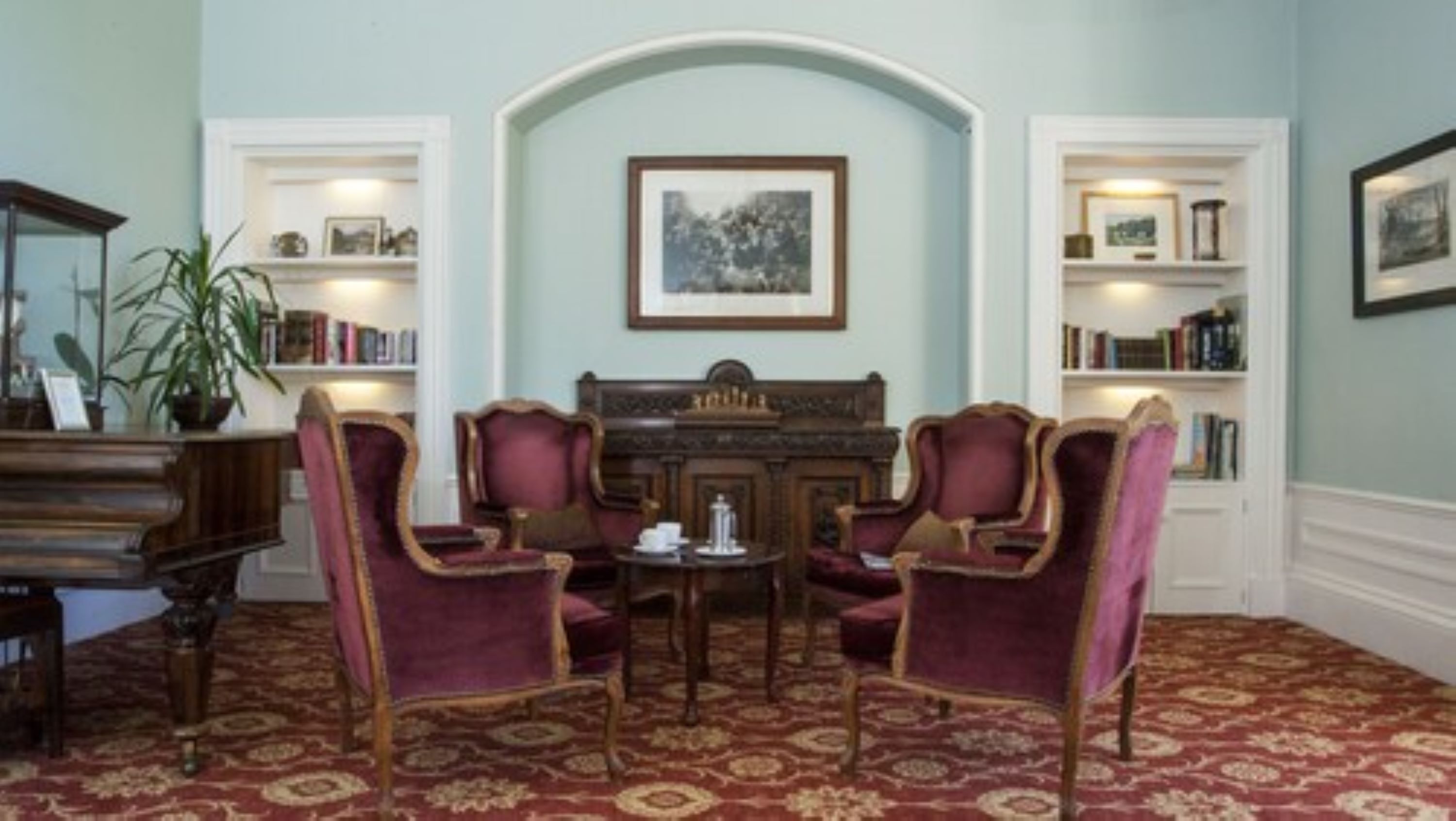A room with a piano, two armchairs, a table with a teapot, and bookshelves.