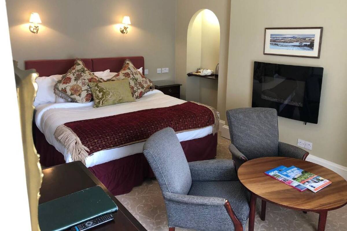 Une chambre d’hôtel avec un lit, deux lampes fixées au mur, une télévision, une table avec des magazines et une chaise.