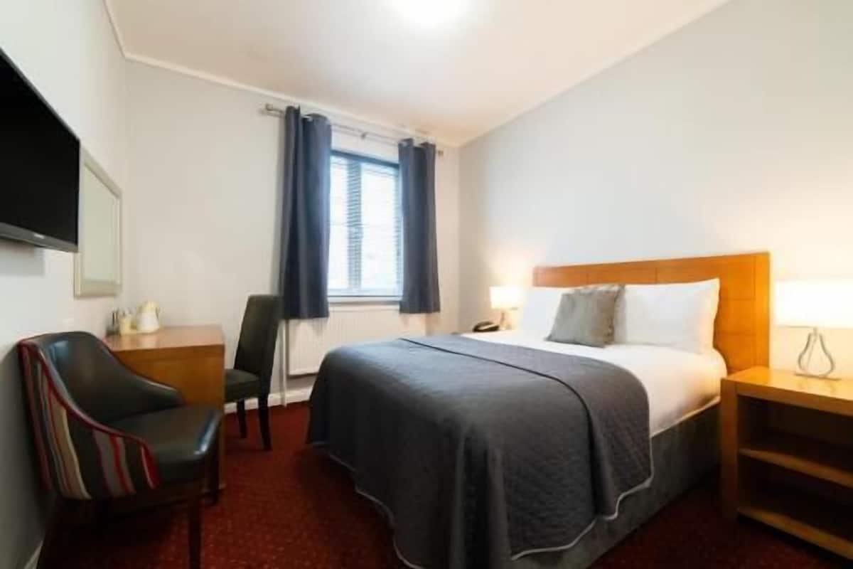 Quarto de hotel com cama, uma cadeira, escrivaninha, televisão e janela com cortinas.