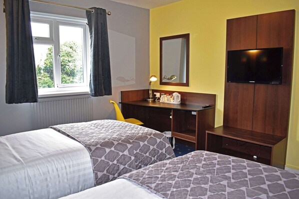 Quarto de hotel com duas camas, uma escrivaninha com cadeira, televisão e janela com cortinas.
