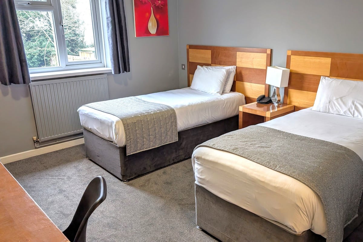 Quarto de hotel com duas camas, uma escrivaninha, uma cadeira, um radiador e uma janela com cortinas.