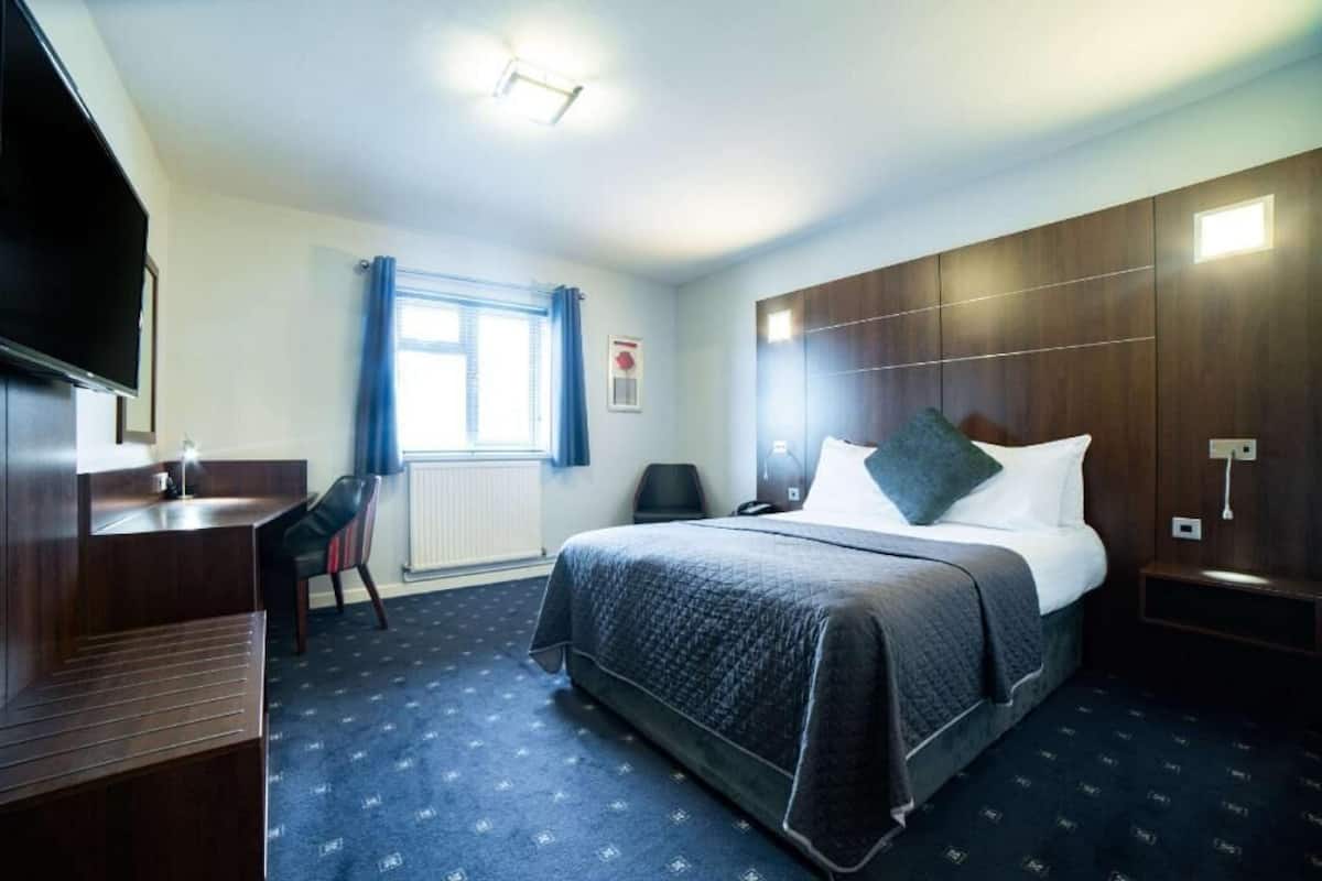 Quarto de hotel com cama, escrivaninha, cadeira, televisão e janela com cortinas.