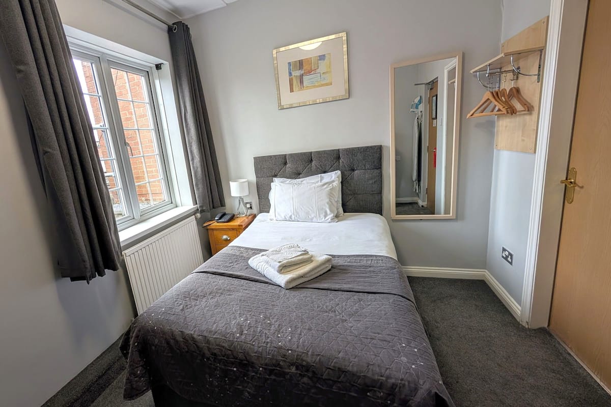 Quarto de hotel com cama, mesa de cabeceira com abajur, janela com cortinas, espelho e cabideiro.