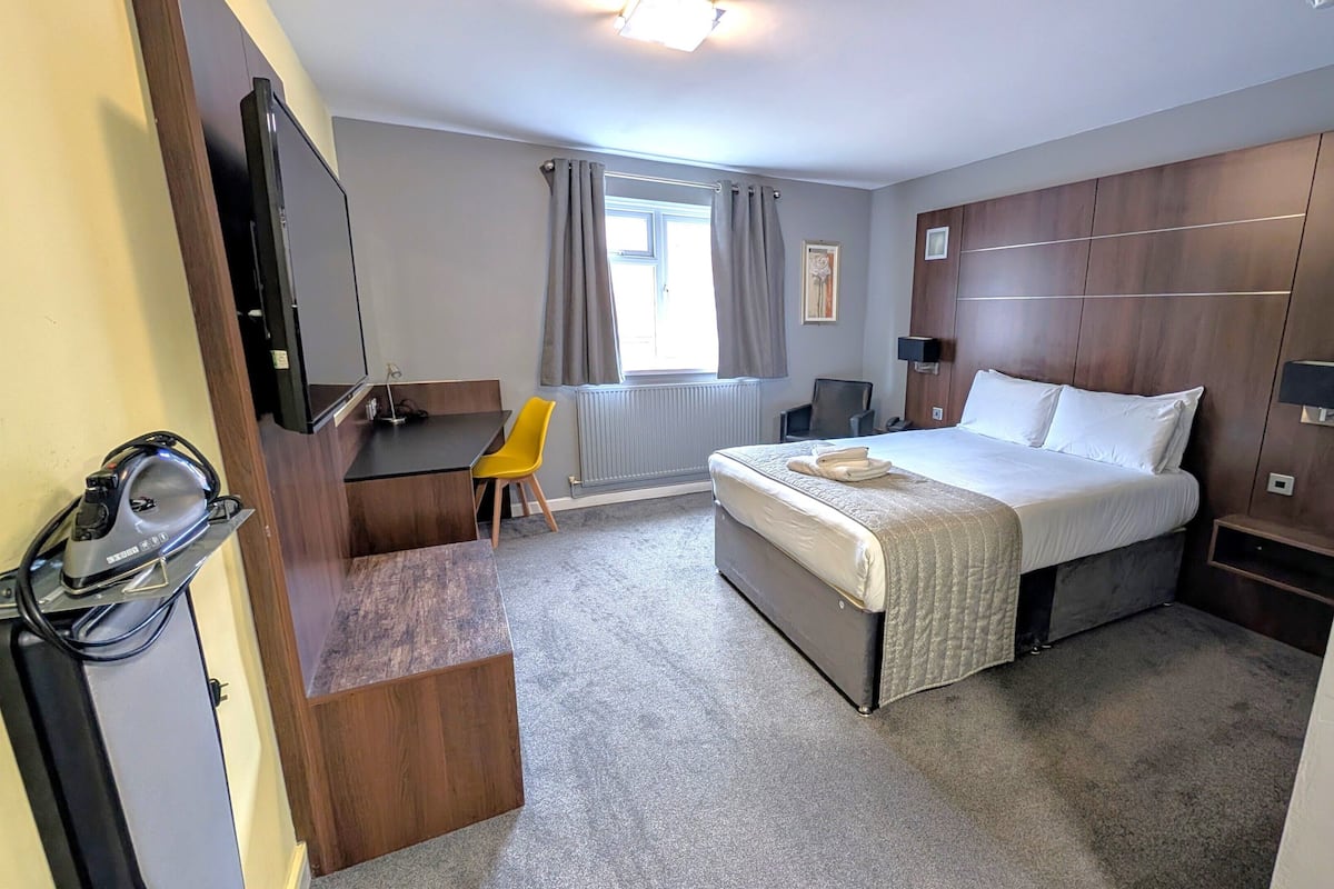 Quarto de hotel com cama, escrivaninha, cadeira, televisão e janela com cortinas.