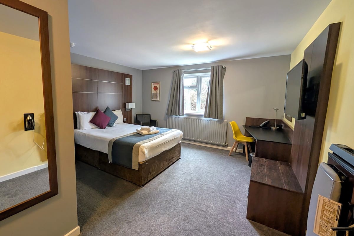 Quarto de hotel com cama, escrivaninha, cadeira, televisão e janela com cortinas.