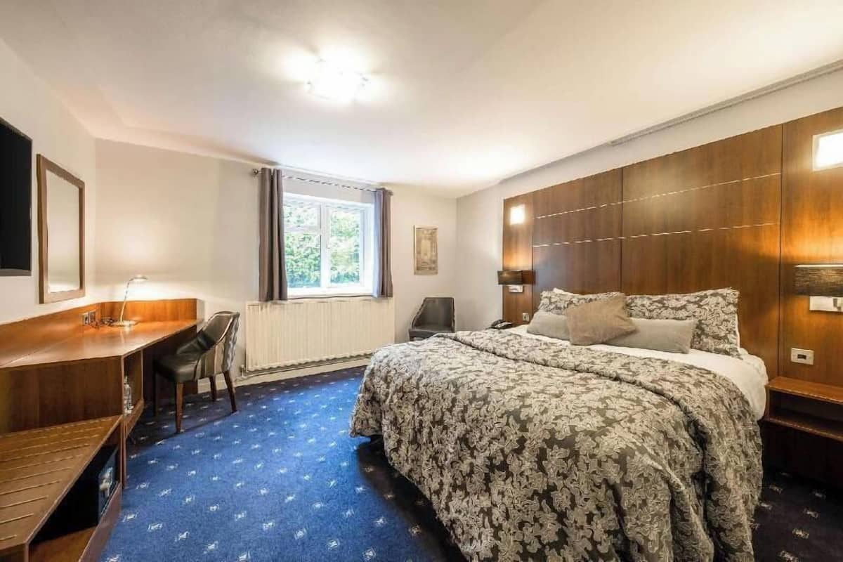 Quarto de hotel com cama, escrivaninha, cadeira, janela com cortinas e radiador.