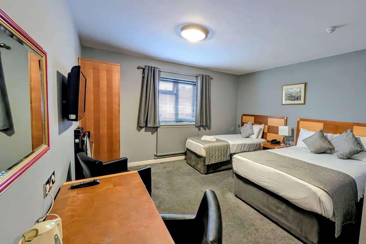 Quarto de hotel com duas camas, uma escrivaninha, uma cadeira, televisão, janela com cortinas e um quadro na parede.