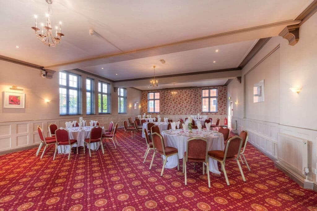 Un salón de banquetes con mesas redondas, alfombra roja estampada y luz natural que entra por grandes ventanales.
