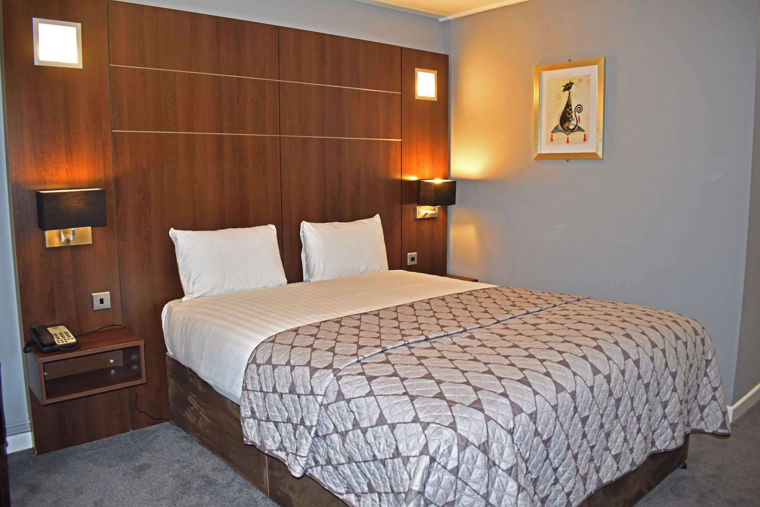 A hotel room with a large bed, wooden headboard, two bedside lamps, a nightstand with a phone, and a framed picture on the wall.