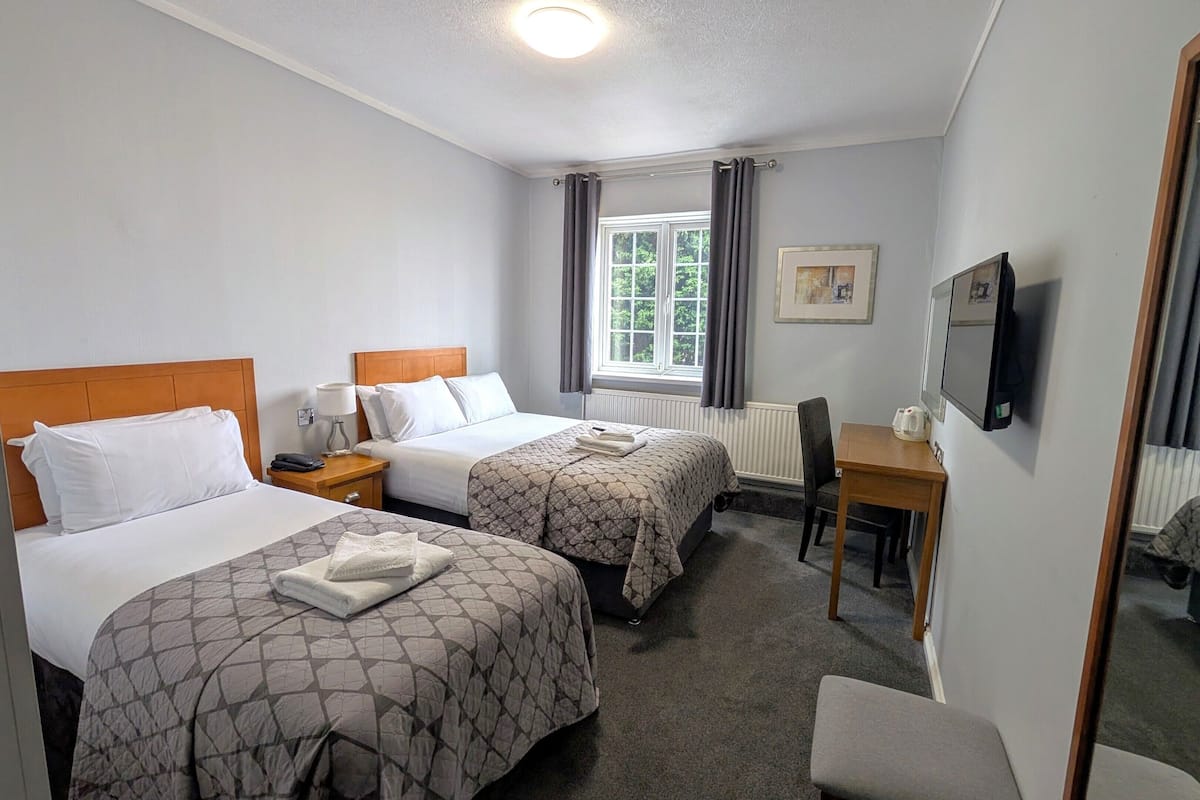Quarto de hotel com duas camas, uma escrivaninha, uma cadeira, uma TV e uma janela com cortinas.