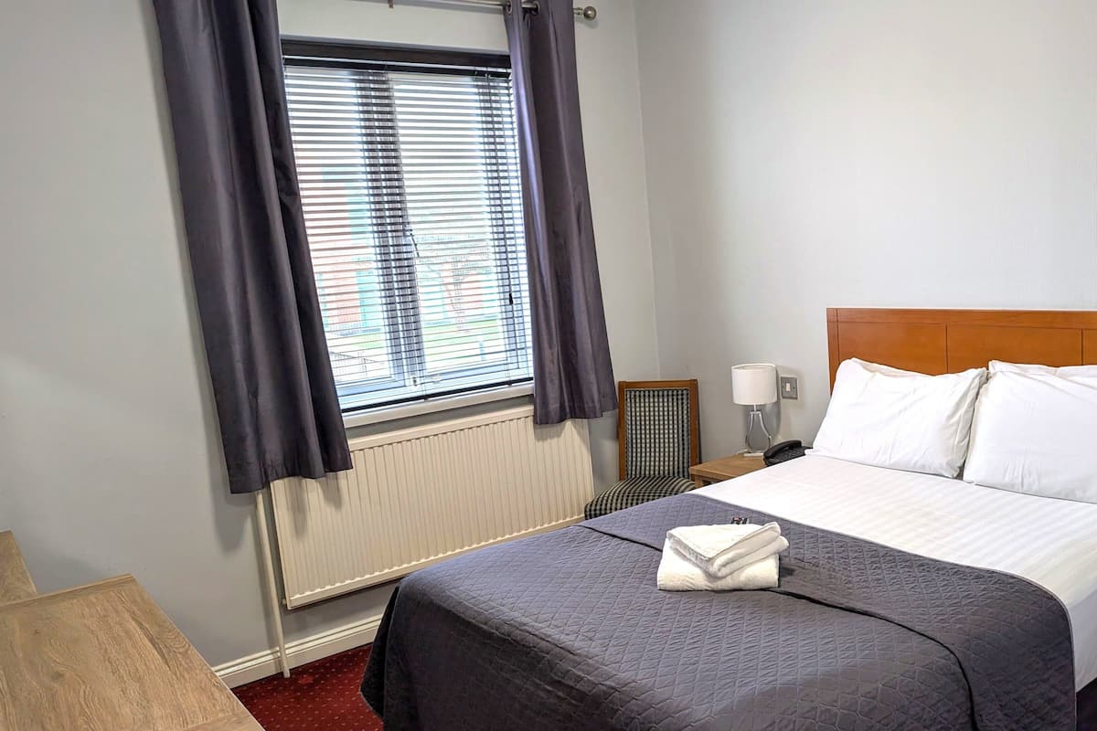 Quarto de hotel com cama, janela com persianas, cadeira, mesa de cabeceira e abajur.