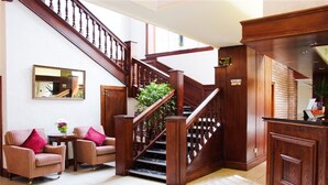 A lobby with a wooden staircase, two armchairs, a small table with a vase, and a reception desk.