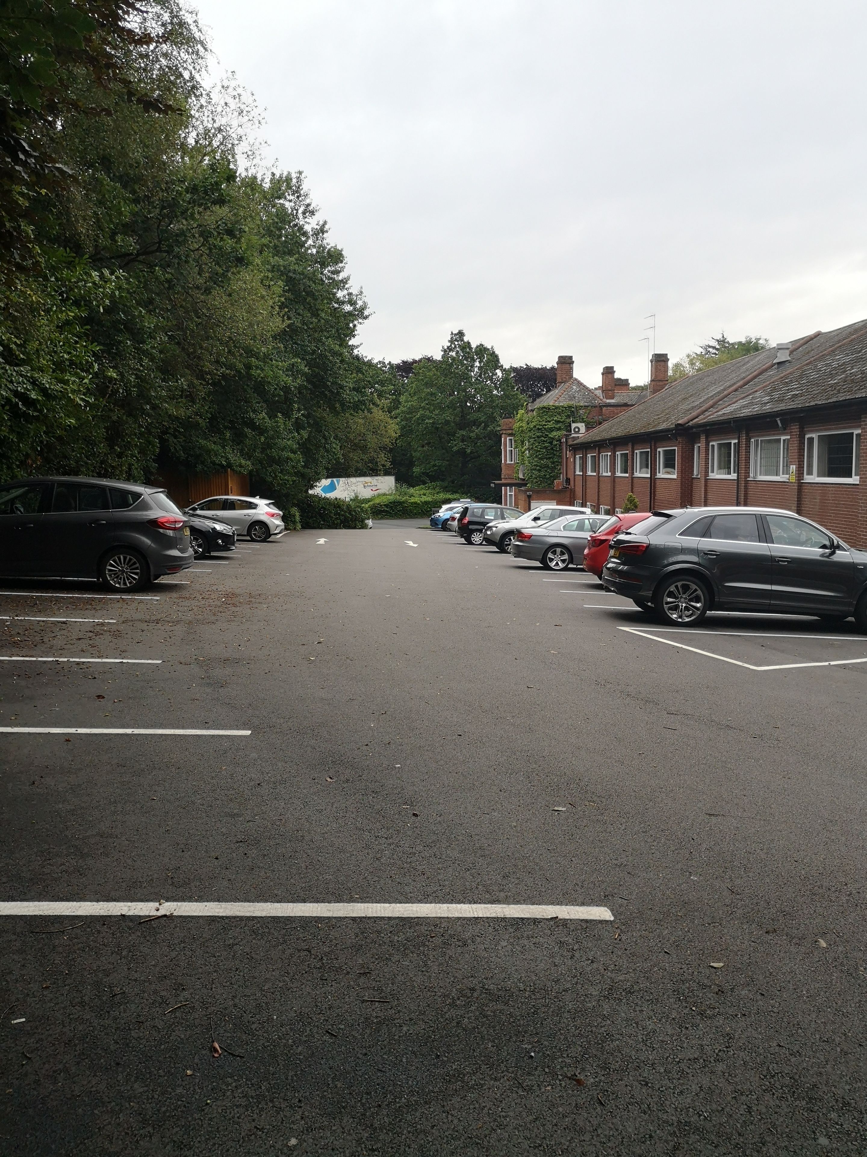 A parking lot with multiple cars parked, surrounded by trees and a building with a tiled roof.