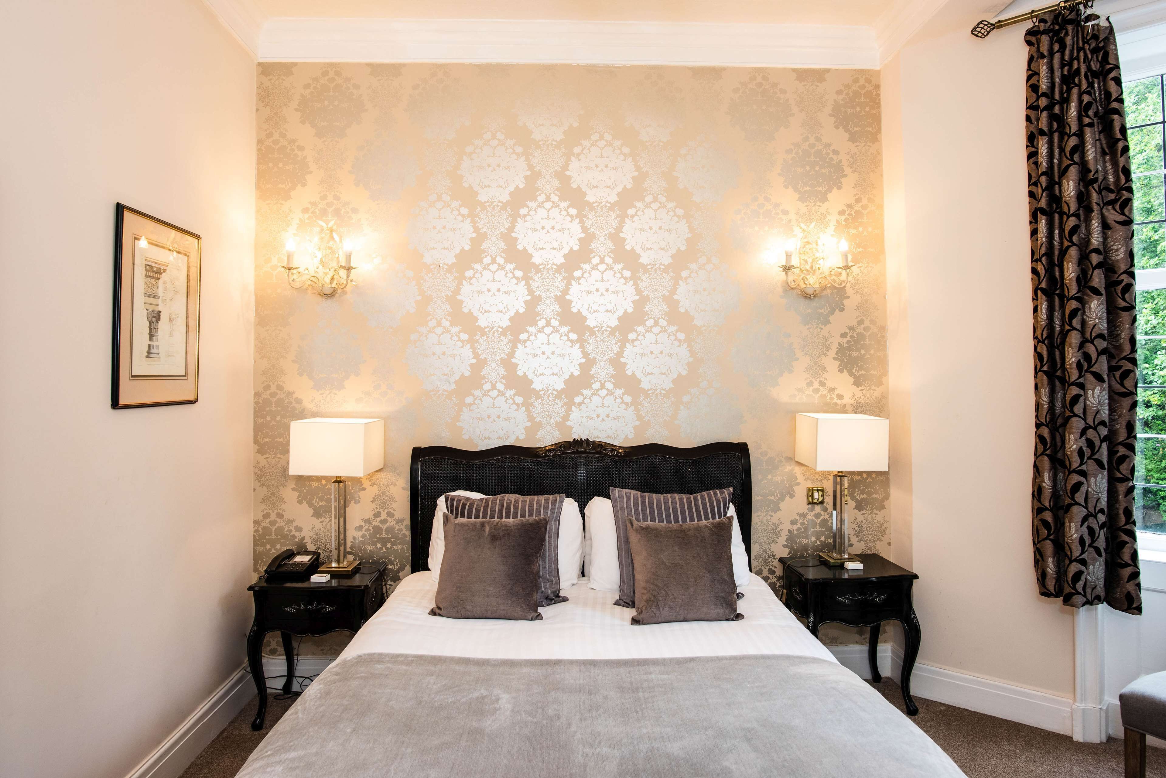 A hotel room with a large bed, two bedside tables with lamps, a framed picture on the wall, and patterned wallpaper.