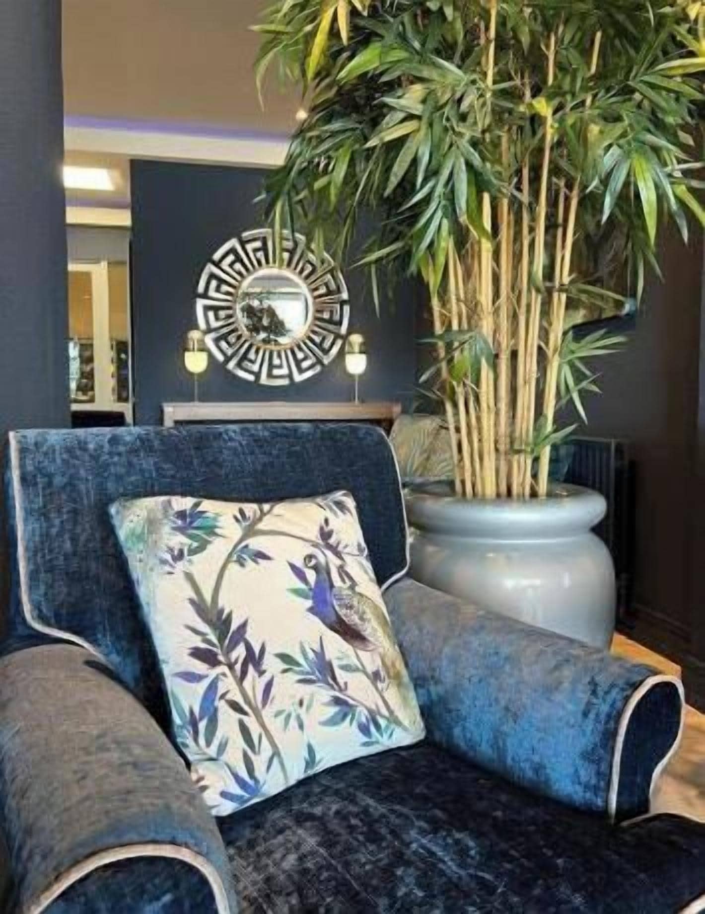 Une pièce comprenant un fauteuil recouvert d’un tissu bleu, un coussin décoratif, une grande plante en pot et une horloge murale.