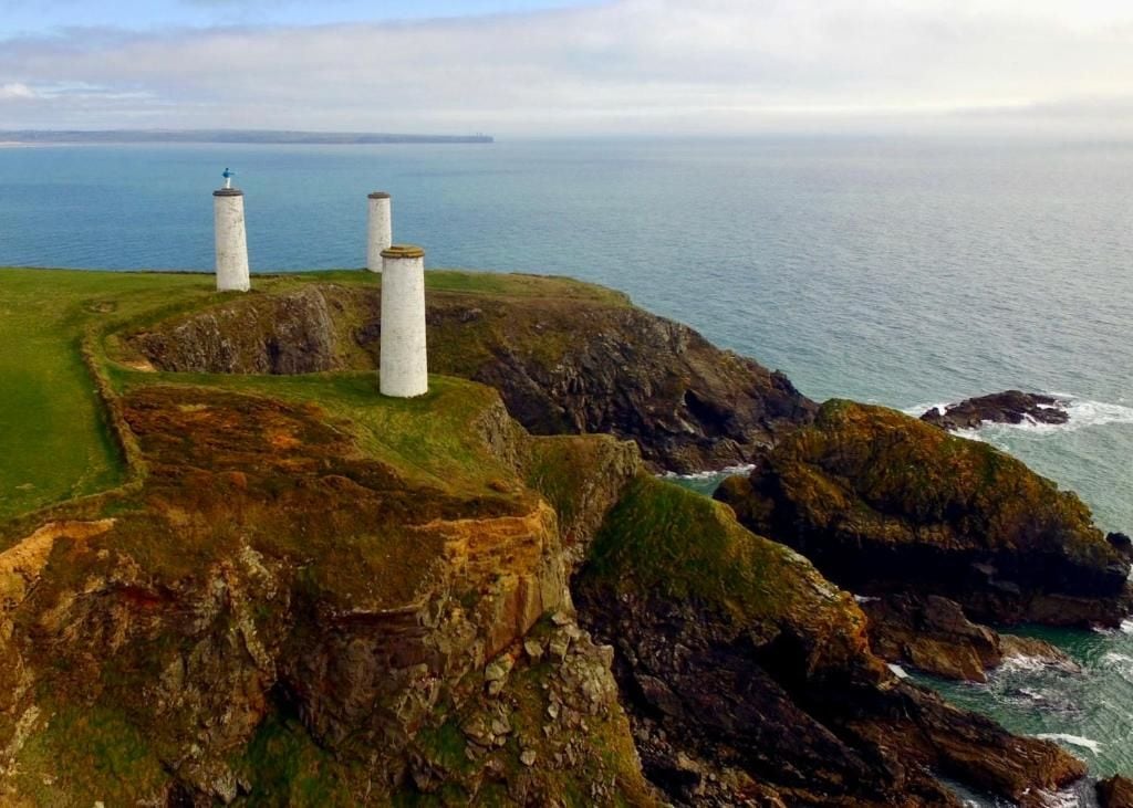 A coastal landscape with three white cylindrical structures, possibly lighthouses, on a rocky outcrop overlooking the sea.