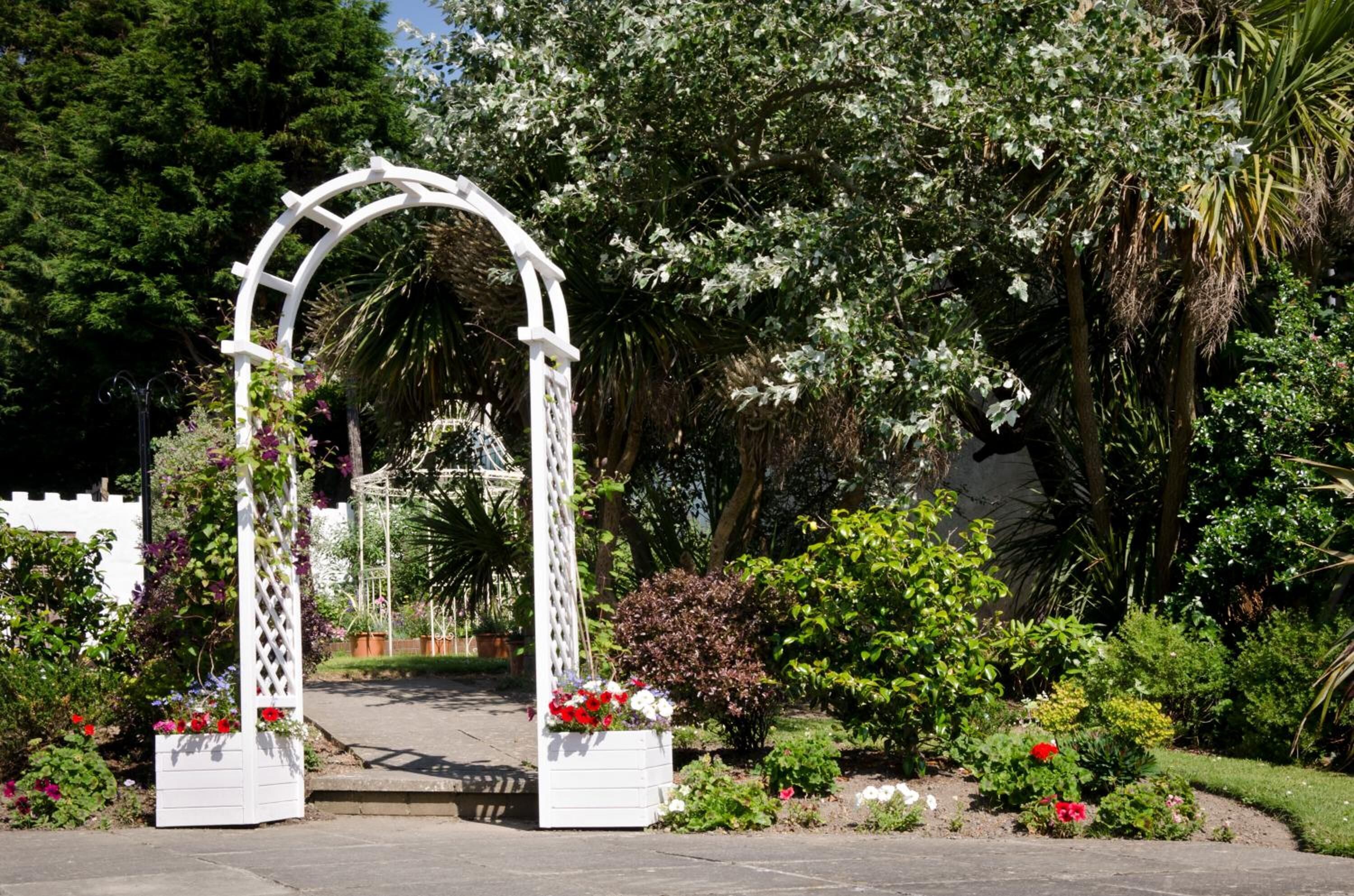 Una entrada de jardín blanca con arco y diseño de celosía, rodeada de abundante follaje y plantas florecientes.
