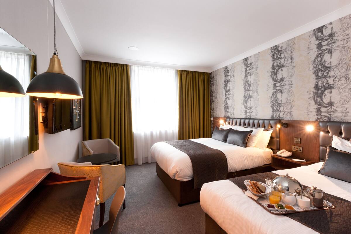A hotel room with a large bed, a smaller bed, a desk with a chair, a dining table with breakfast items, and a large mirror.