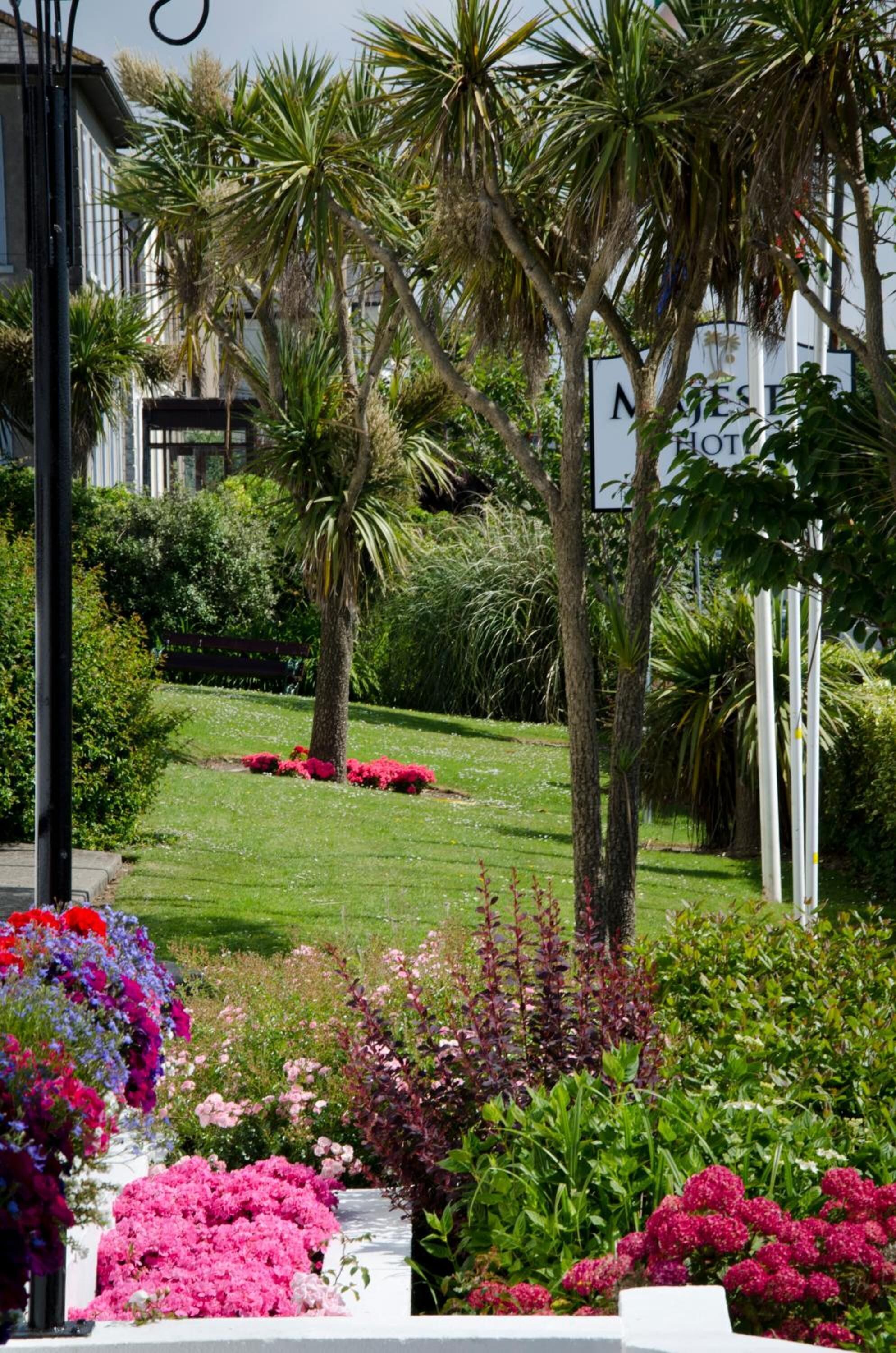 A well-maintained garden with various flowering plants and palm trees, a bench, and a building with a sign that reads "Mansion Hotel".
