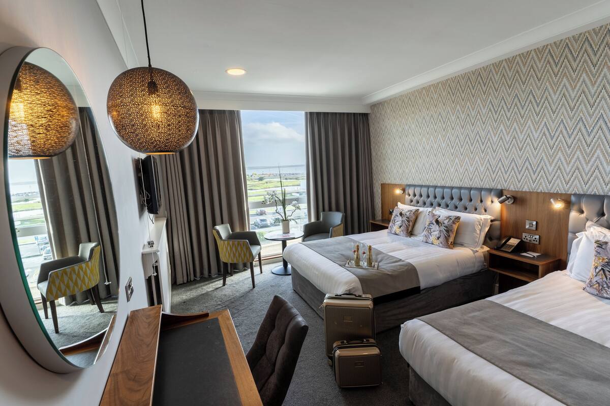 A hotel room with two beds, a desk, a chair, a mirror, and a view of the outside.