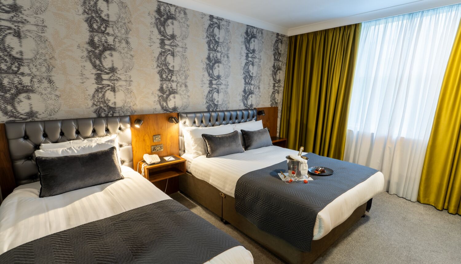 A hotel room with two beds, a headboard with a light fixture, a nightstand with a phone and a glass of water, and a window with yellow curtains.