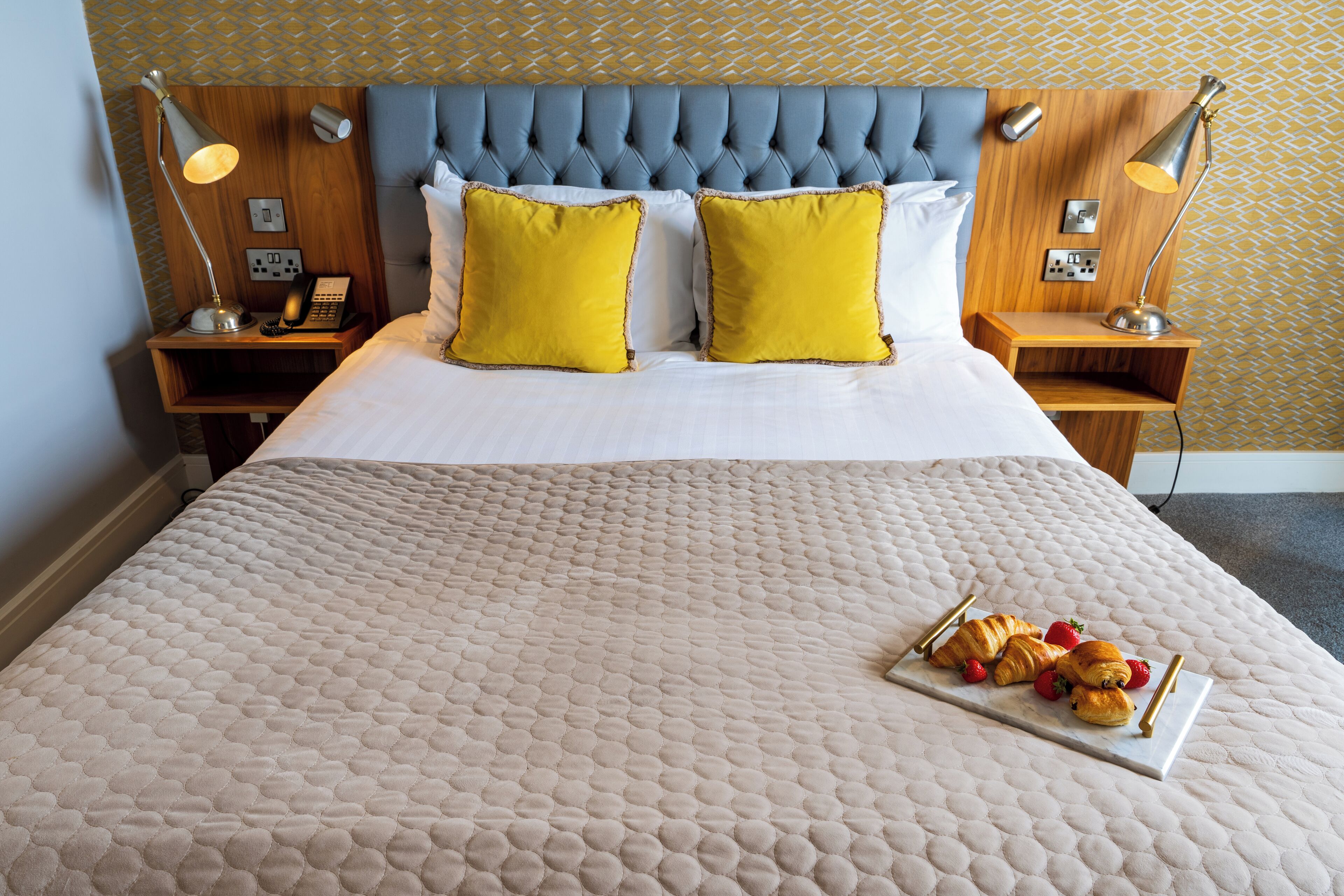 Un lit bien fait, recouvert d’une courtepointe beige, avec deux coussins jaune vif et un plateau contenant des croissants et des fraises posé sur le lit.