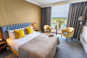 Une chambre d’hôtel avec un grand lit, deux fauteuils, une table ornée d’un vase de fleurs et une vue sur le paysage.