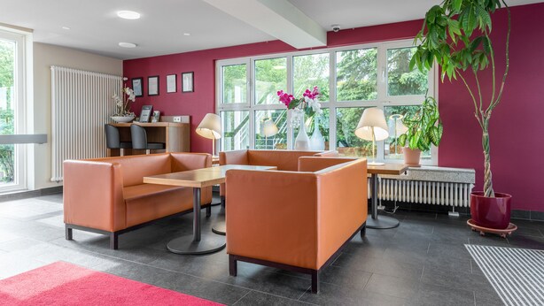 Un área de espera moderna con sofás naranja, una mesa de madera y plantas en macetas.