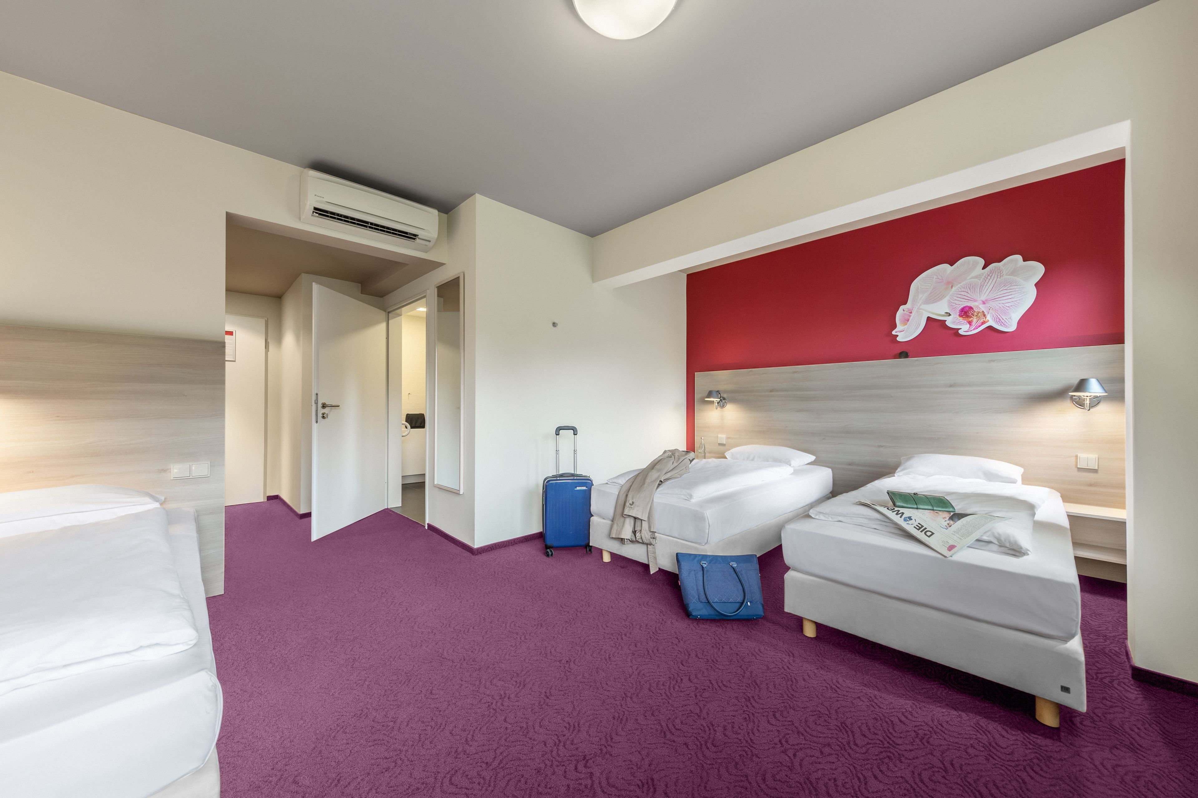 Ein Hotelzimmer mit zwei Betten, einem violetten Teppich, einer roten Akzentwand und einem blauen Koffer.