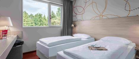 A hotel room with two beds, a desk, a lamp, and a window with a view of trees.