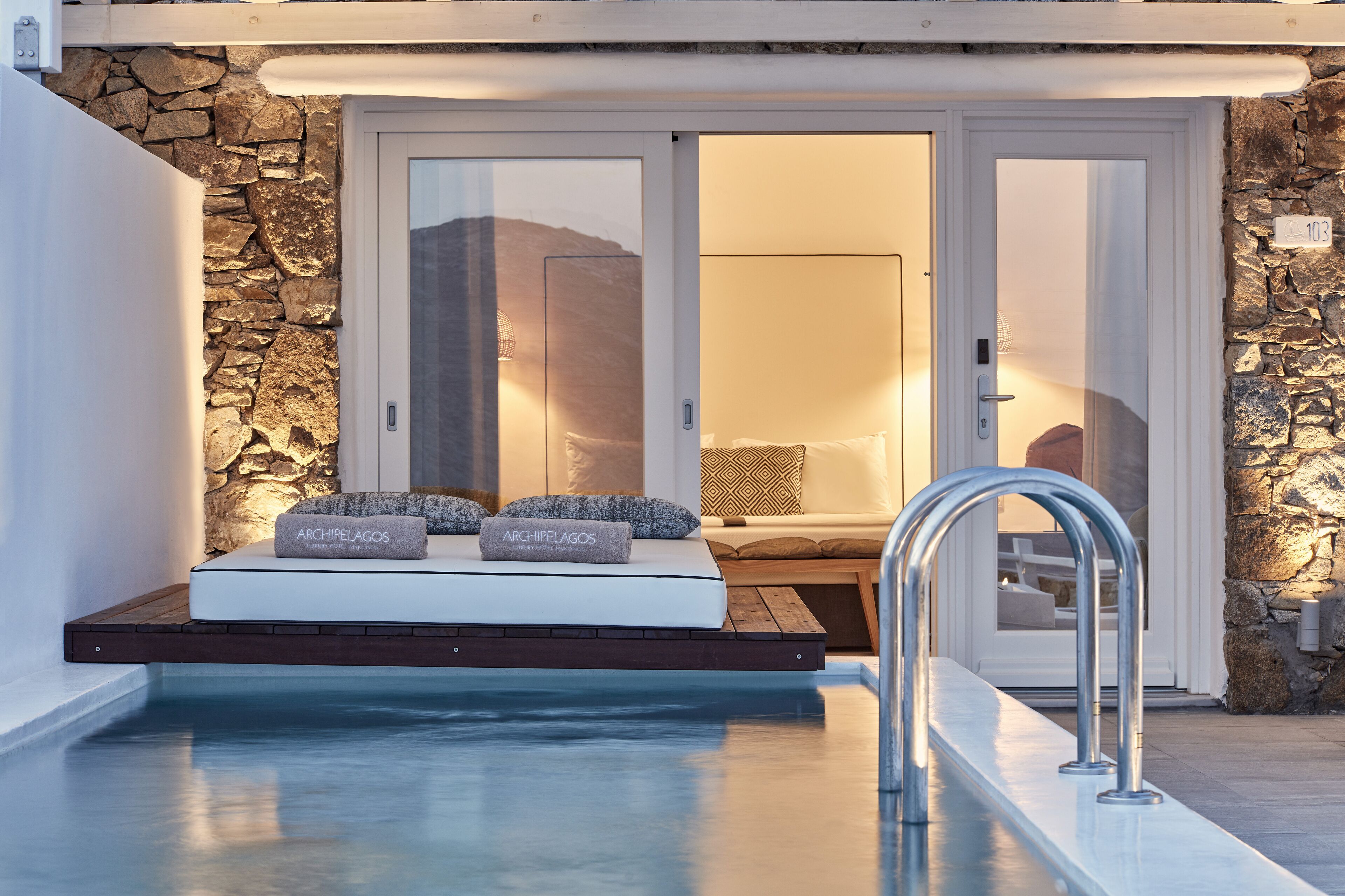 A poolside area with a stone wall, a bed with pillows, and a metal handrail.