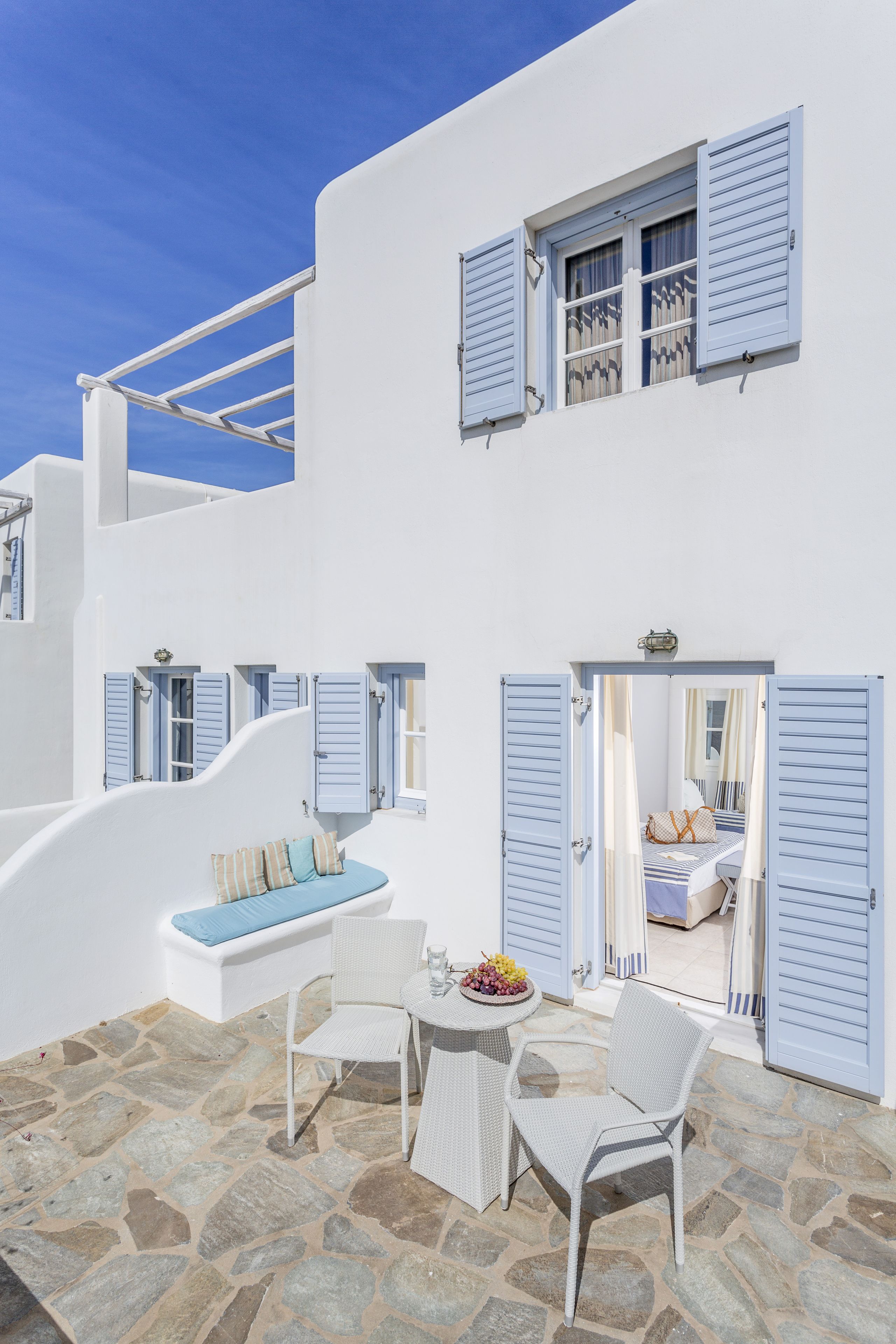 Un edificio blanco con contraventanas azules, suelo de piedra y un pequeño espacio al aire libre con mesa y sillas.