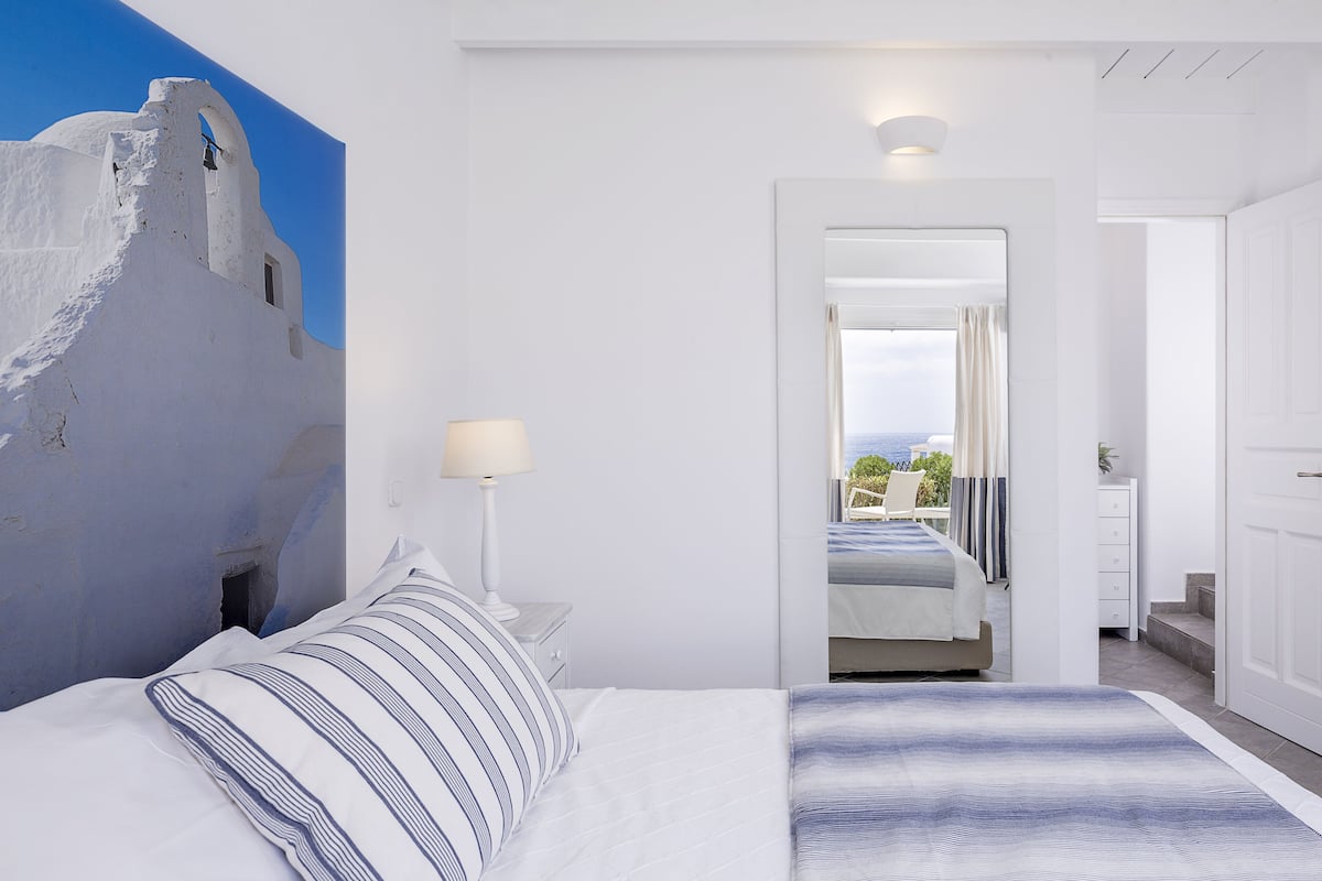 Una habitación de hotel con cama, mesita de noche, lámpara y vista al mar a través de una puerta de balcón.