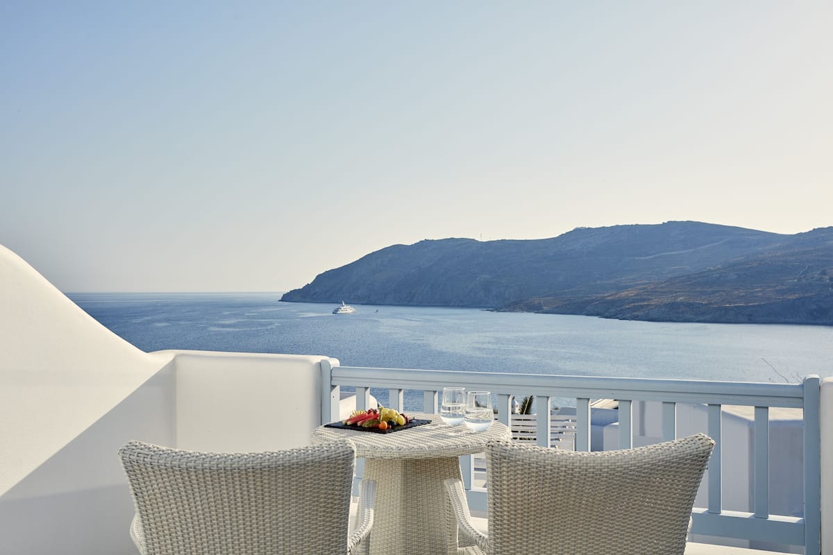 Un balcón con sillas de mimbre y una mesa, con vista a un mar tranquilo y montañas lejanas.