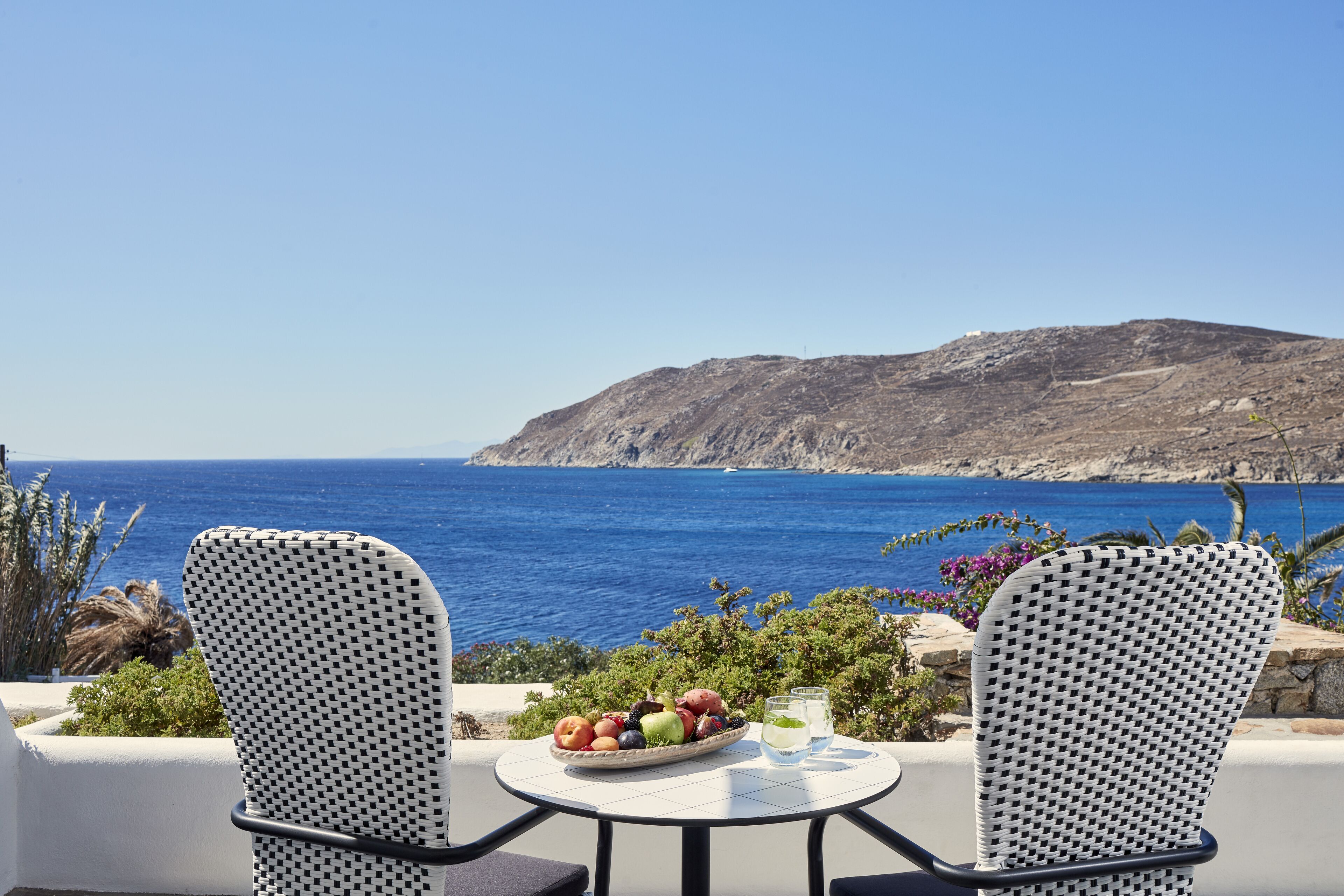 A table with a fruit bowl and glasses, overlooking a coastal view with a rocky hill.