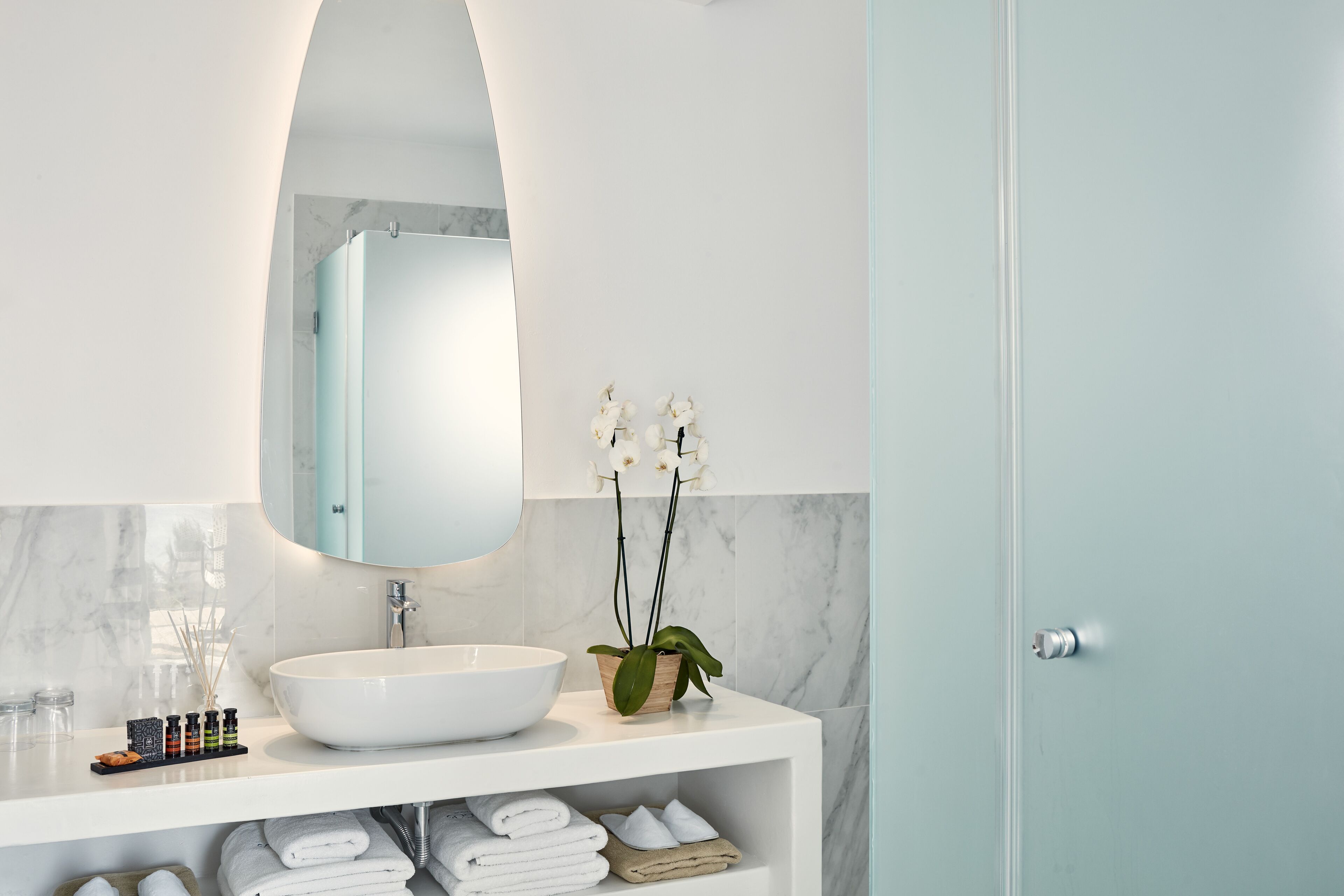 Une salle de bain avec un lavabo blanc, un miroir et une étagère où sont posées des serviettes et une petite plante.
